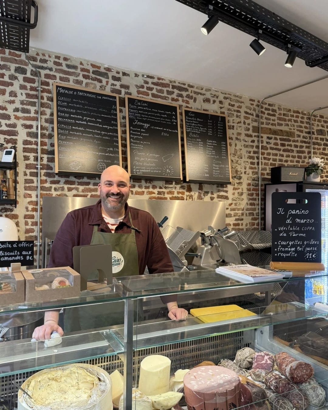 Joachim Tordi derrière son comptoir, devant la vitrine de charcuteries et de fromages, sous les ardoises de la carte